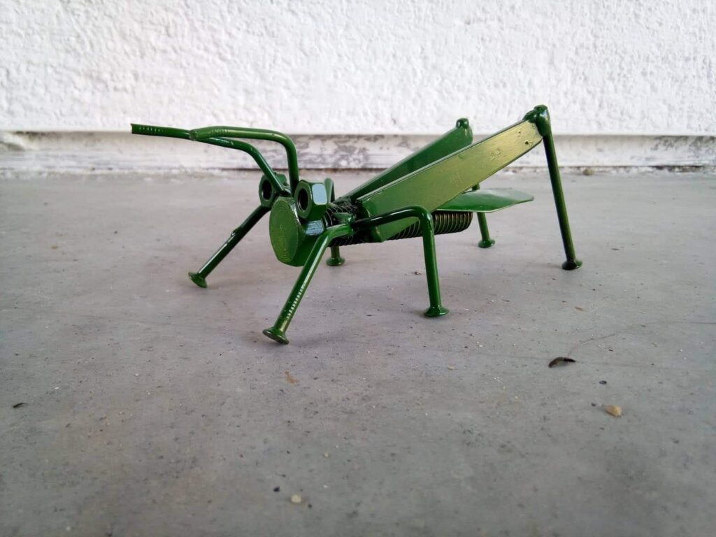 Ručně vyráběná kovová socha kobylky ze šroubu, dvou matek jako očí a hřebíků jako nohou, s komaxitovým nátěrem.